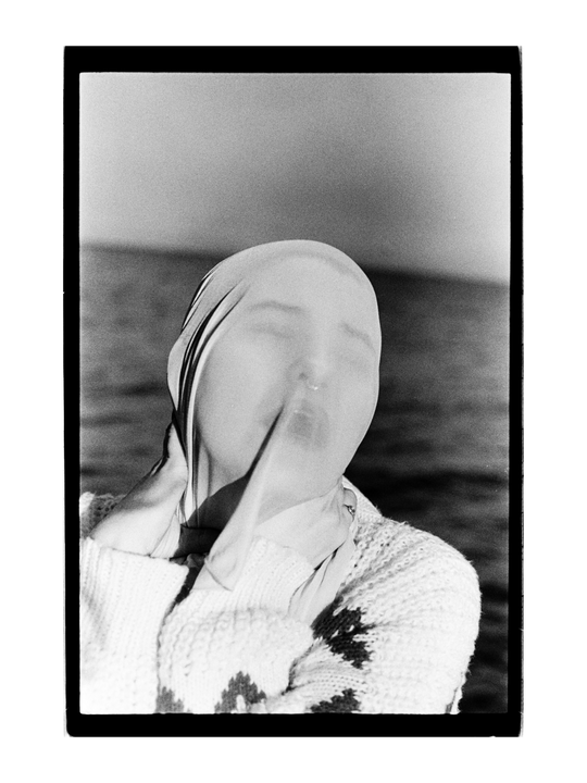 A person with a stocking over their head is wearing a knitted sweater and standing in front of water.