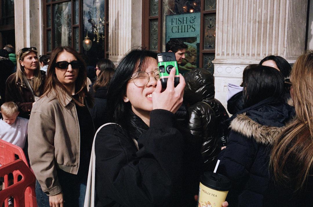 A woman in glasses holds a green disposable camera to her face, surrounded by other people on a street.