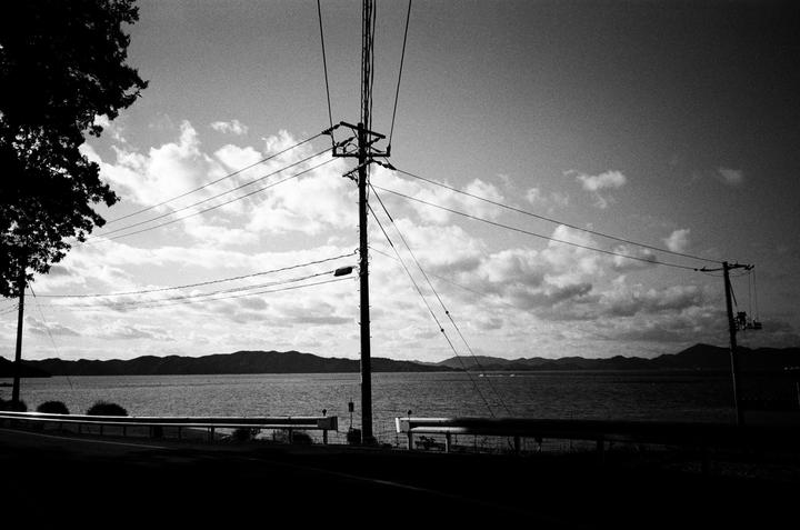 A black and white landscape shows a body of water with mountains in the distance, a utility pole with many wires, and a tree on the left.